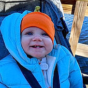 Autumn is registered to the contest to win money with this photo: baby, child, smiling, blue_jacket, orange_hat, stroller, outdoor, water, wooden_dock, sunny, happy, face, person, winter_clothing, bundle_up, seatbelt, nature, daylight, cute, portrait