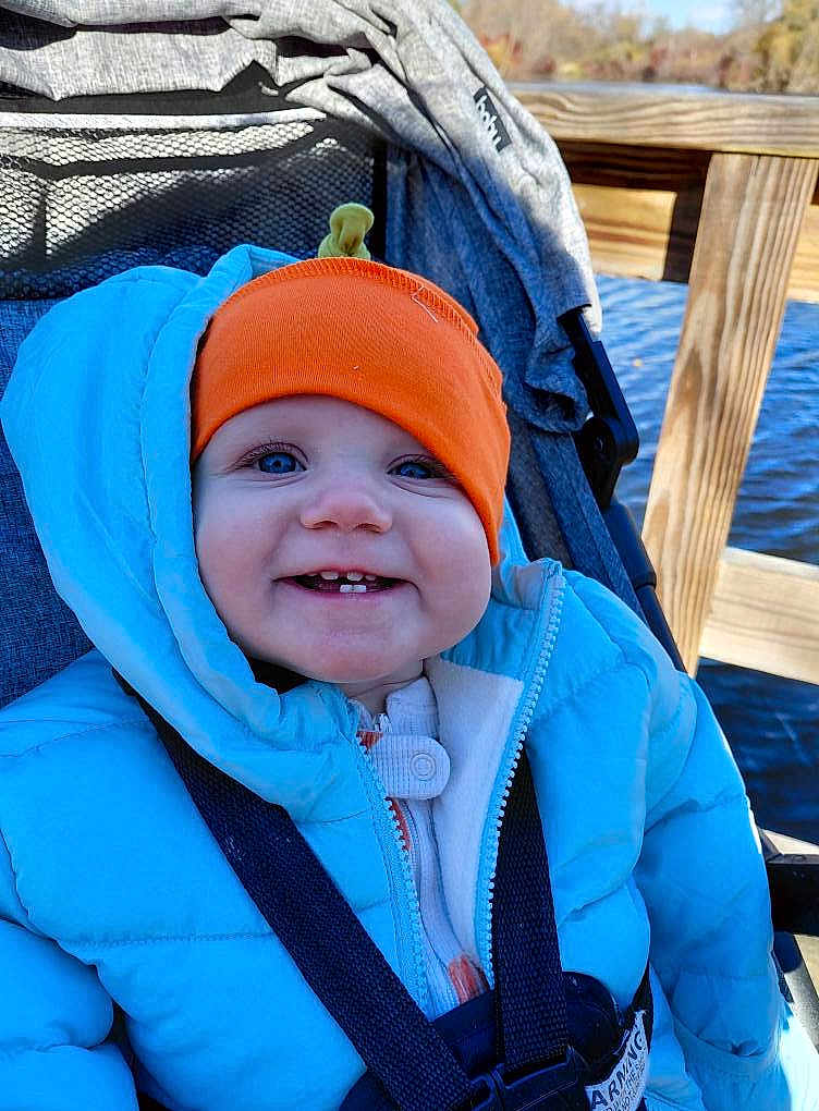 Autumn is registered to the contest to win money with this photo: baby, child, smiling, blue_jacket, orange_hat, stroller, outdoor, water, wooden_dock, sunny, happy, face, person, winter_clothing, bundle_up, seatbelt, nature, daylight, cute, portrait