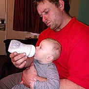 Adam is registered to the contest to win money with this photo: man, baby, feeding, bottle, indoor, person, clothing, cap, red_shirt, striped_shirt, baby_clothes, holding, care, parenting, room, curtain, window, furniture, milk, focus