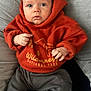 baby, infant, hoodie, orange_clothing, grey_pants, checkered_socks, cushion, soft_fabric, indoor, child, portrait, cute, cozy, sitting, hands, face, eyes, expression, comfortable, casual_clothing