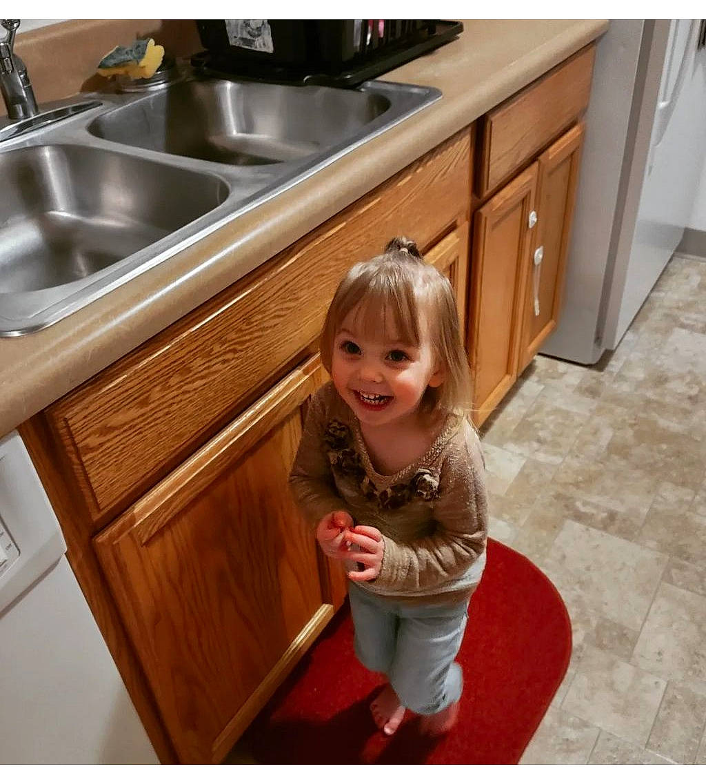Luna is registered to the contest to win money with this photo: baby_toddler_clothing, cabinetry, countertop, floor, flooring, hardwood, joy, kitchen, kitchen_sink, person, photograph, plumbing_fixture, room, sink, smile, snapshot, tap, toddler, varnish, white