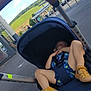 child, stroller, sleeping, outdoor, parking_lot, shoes, sky, clouds, hill, trees, person, summer_clothing, casual, metal_poles, asphalt, daylight, relaxing, vacation, urban, nature