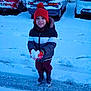 child, snow, winter_clothing, hat, gloves, jacket, boots, parking_lot, car, snow_covered, outdoor, smiling, holding_snow, cold_weather, snowy_ground, red_hat, casual, standing, daylight, playful