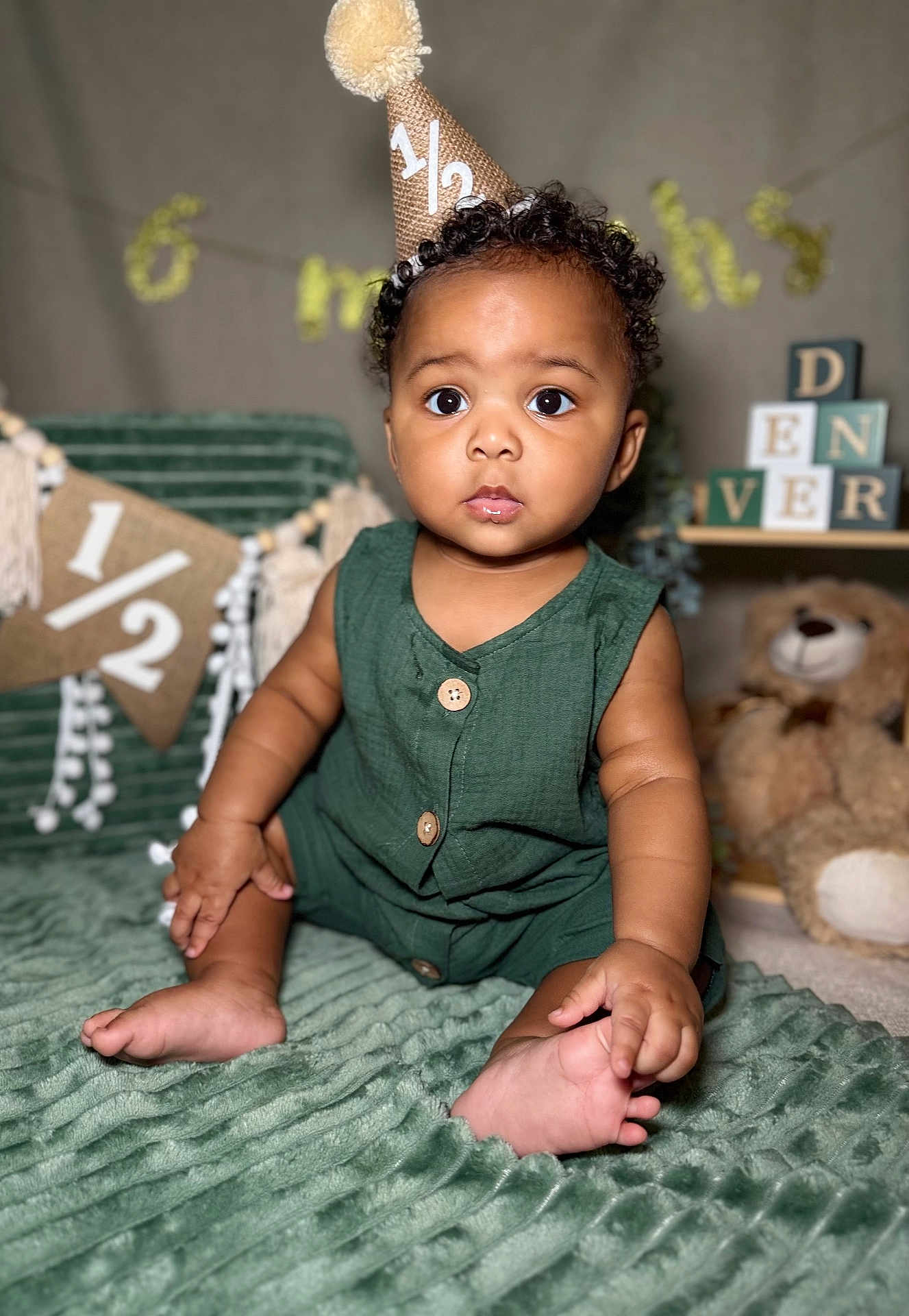 Denver is registered to the contest to win money with this photo: baby, child, party_hat, green_outfit, sitting, blanket, teddy_bear, decorations, milestone, curious, wide_eyes, cute, indoor, portrait, celebration, soft_texture, buttoned_outfit, fuzzy, infant, birthday