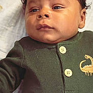 Titus is registered to the contest to win money with this photo: baby, infant, face, portrait, clothing, green_outfit, button, dinosaur_embroidery, lying_down, blanket, soft_texture, young_child, cute, indoors, child, person, newborn, head, skin, eyes