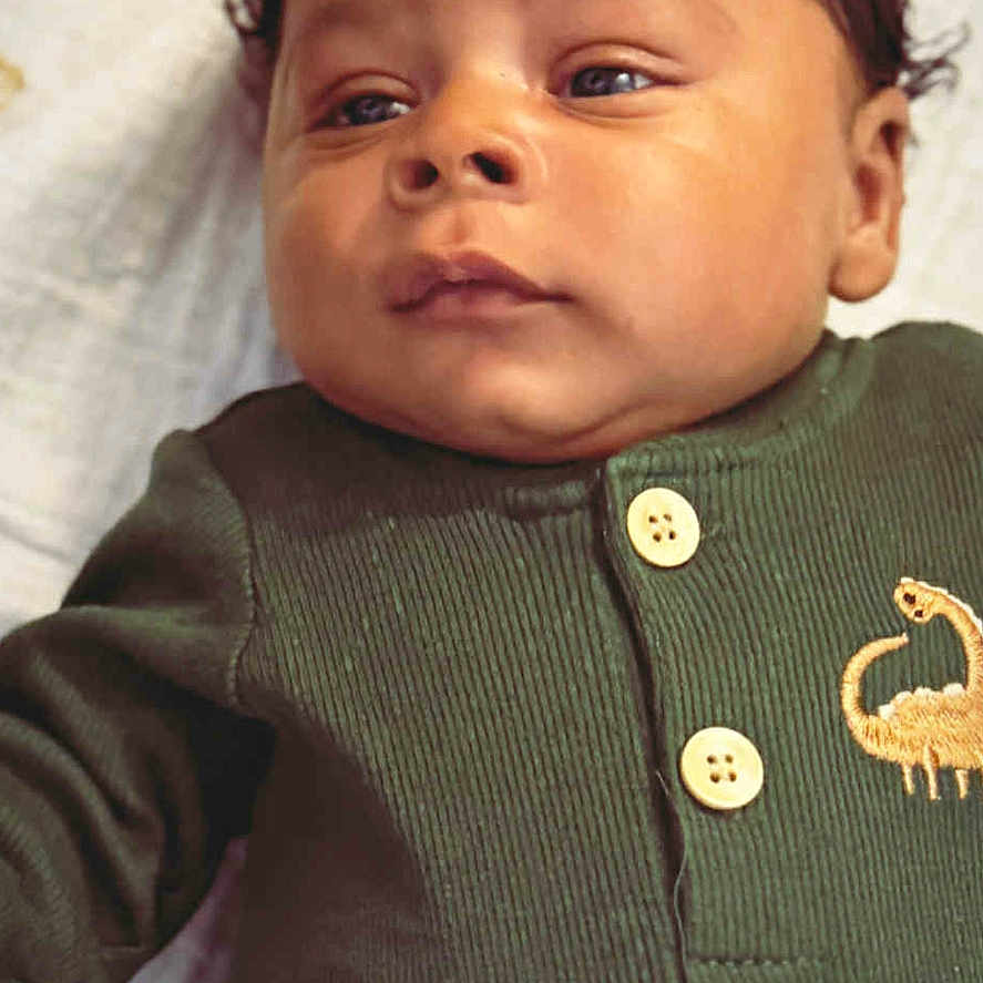 Titus is registered to the contest to win money with this photo: baby, blanket, button, child, clothing, cute, dinosaur_embroidery, eyes, face, green_outfit, head, indoors, infant, lying_down, newborn, person, portrait, skin, soft_texture, young_child