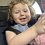 child, toddler, curly_hair, smiling, car_seat, seat_belt, stuffed_animal, pink, toy, happy, indoor, vehicle, person, face, clothing, button, arm, window, light, hair