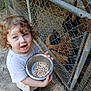 child, toddler, curly_hair, barefoot, bowl, birdseed, chicken_feed, dirt, face, wire_fence, chicken_coop, chickens, outdoor, farm, feeding, animal, person, nature, daylight, curious