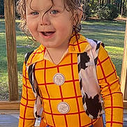 Johnathan is registered to the contest to win money with this photo: child, toddler, costume, woody, toy_story, outdoor, porch, wooden_rail, forest, greenery, smiling, happy, curly_hair, yellow_shirt, blue_pants, belt, vest, playful, sunlight, portrait