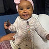 baby, child, close_up, couch, crumbs, family_photo, floral_outfit, hand, happy, headband, indoor, infant, onesie, pink_bow, portrait, seated, smiling, snack, suspenders, toy