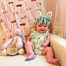 baby, bunny_ears, stuffed_animal, pink, striped_clothing, chair, blanket, easter, sign, smiling, indoors, cute, child, plush_toy, headband, pastel_colors, comfort, decor, holiday, portrait