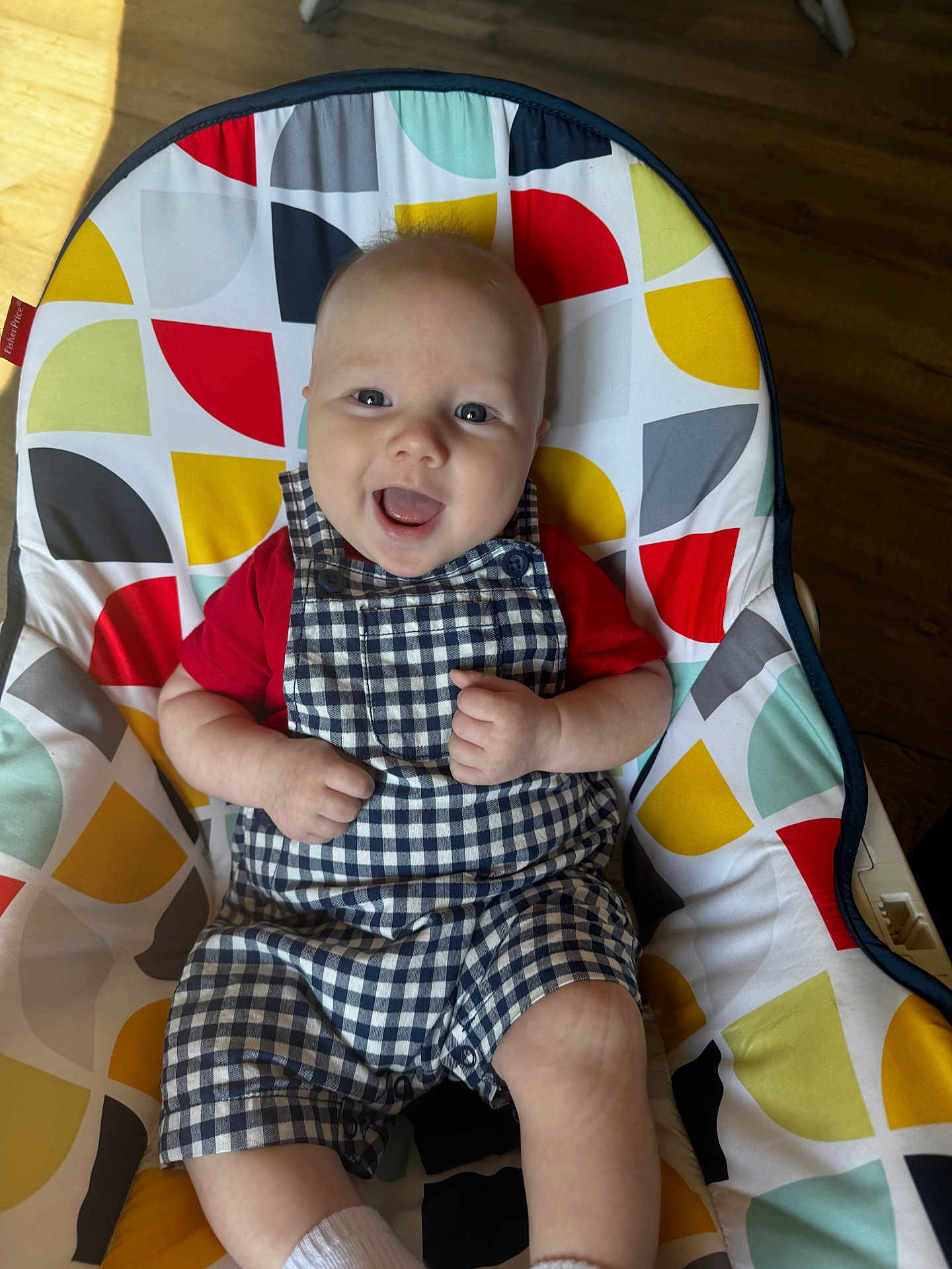 Hudson is registered to the contest to win money with this photo: baby, child, smile, happy, overalls, red_shirt, checkered_pattern, baby_chair, colorful, patterned, indoor, wooden_floor, cute, infant, sitting, portrait, clothing, face, hands, feet