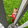 child, toddler, red_dress, smiling, greenery, grass, outdoor, wooden_fence, wood, nature, playful, happy, person, portrait, summer, daylight, curious, candid, standing, garden