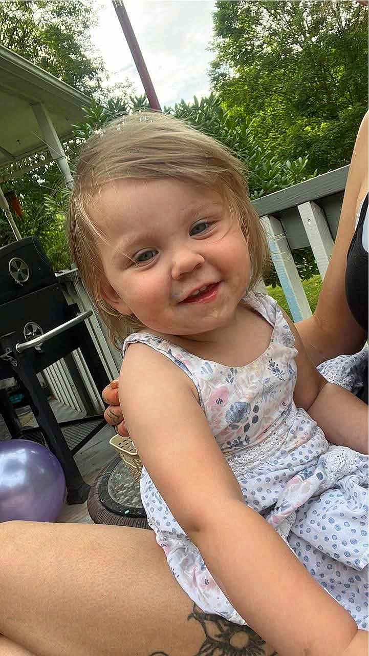 Liala is registered to the contest to win money with this photo: toddler, child, smiling, outdoor, porch, dress, floral_pattern, greenery, balloon, grill, summer, happy, person, nature, casual, tattoo, arm, daylight, family, relaxation