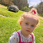 Karlene is registered to the contest to win money with this photo: child, toddler, girl, grass, outdoor, vintage_truck, trees, sunny_day, greenery, curly_hair, pink_bow, casual_clothing, portrait, nature, daylight, field, expression, playing, young_child, relaxed