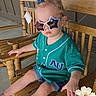 bow, casual_clothing, child, cute, decorative_flowers, fashion, flowers, footwear, green_shirt, hair_accessory, outdoor, porch, rocking_chair, serious_expression, shorts, star_shaped, summer, sunglasses, toddler, wooden_chair