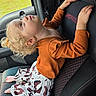 baby, blonde_hair, car_interior, car_seat, casual_clothing, child, curious_expression, curly_hair, cute, feet, green_background, hands, indoors, orange_top, patterned_pants, seatbelt, side_view, toddler, window, young_child