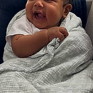 Noah is registered to the contest to win money with this photo: baby, child, blanket, couch, smile, happy, indoors, infant, person, soft_texture, wrapped, seated, clothing, hand, face, cute, joyful, dark_hair, expression, comfort