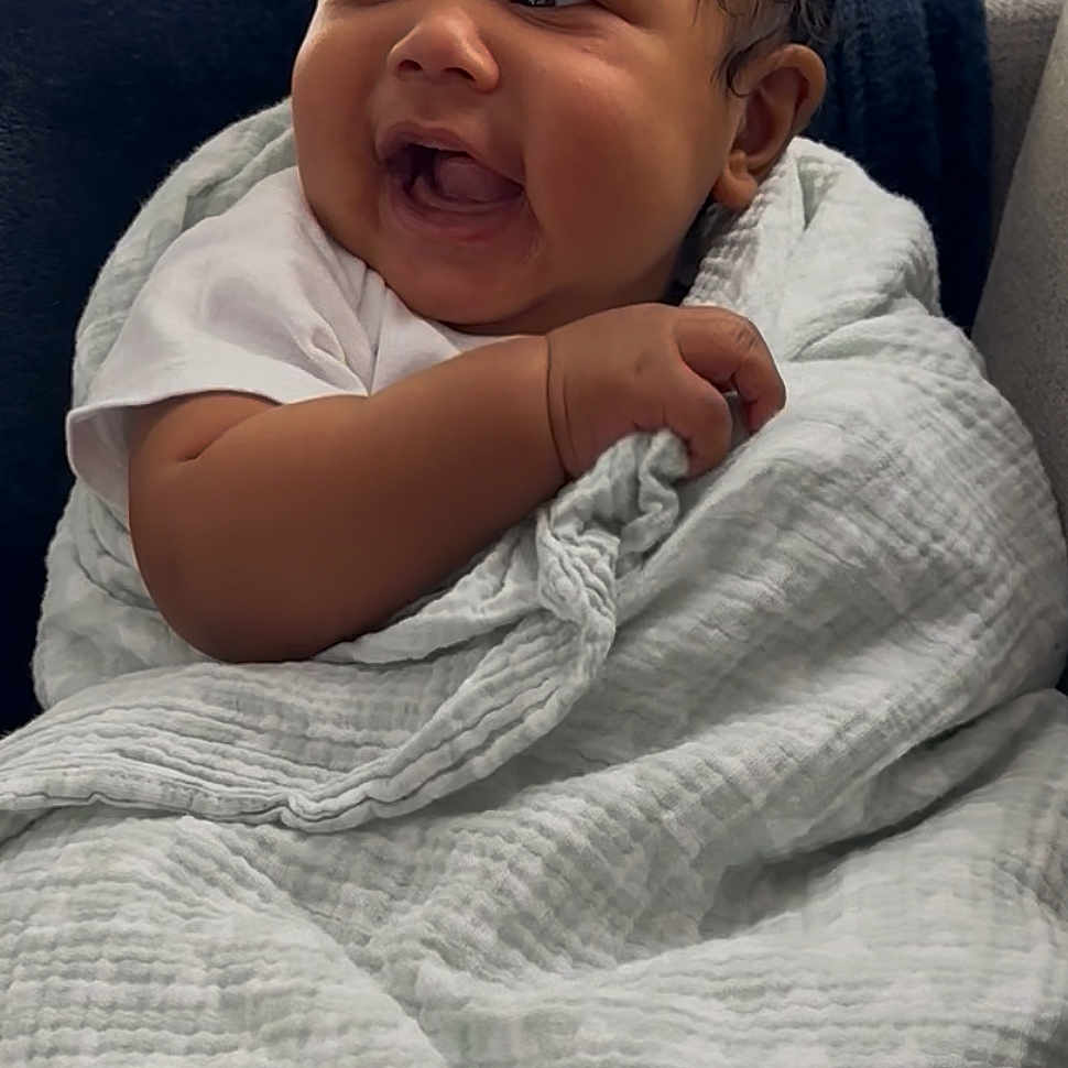 Noah is registered to the contest to win money with this photo: baby, blanket, child, clothing, comfort, couch, cute, dark_hair, expression, face, hand, happy, indoors, infant, joyful, person, seated, smile, soft_texture, wrapped