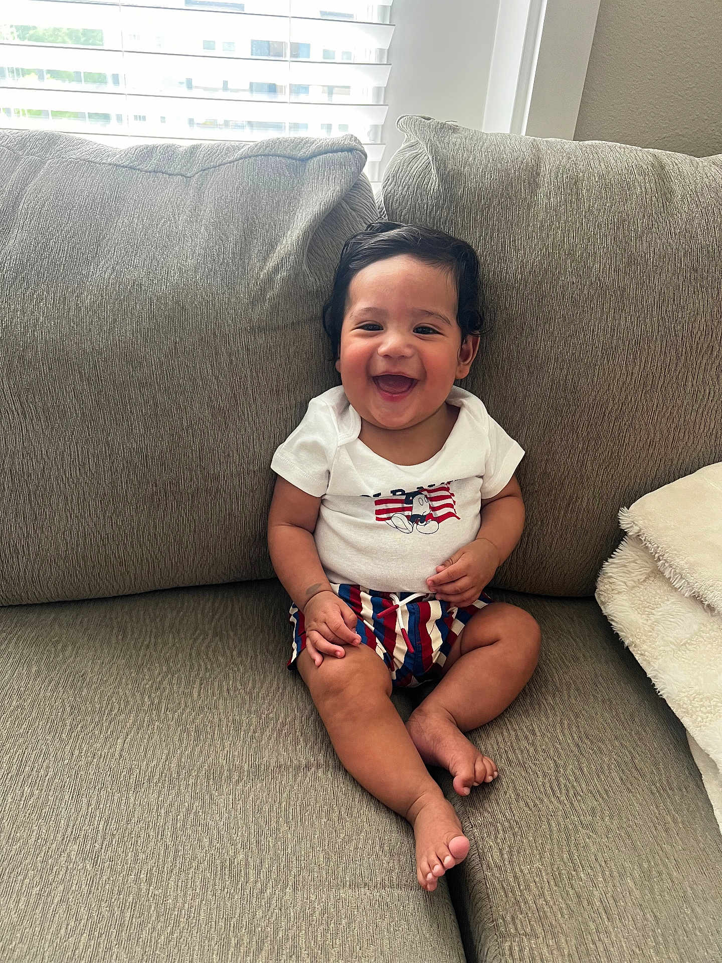 Tommy is registered to the contest to win money with this photo: baby, blanket, child, clothing, couch, curly_hair, cute, face, foot, hand, happy, home, indoor, infant, person, shorts, sitting, smiling, tshirt, window
