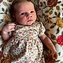 baby, infant, child, floral_dress, blanket, pattern, face, hand, skin, person, lying_down, cute, young, indoors, portrait, expression, newborn, soft, comfortable, clothing