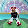 toddler, child, sitting, red_bows, braided_hair, black_tshirt, jeans, red_black_sneakers, playground, colorful_background, starburst_pattern, outdoor, happy, smiling, young_child, fashionable, portrait, fun, green_mound, casual_clothing