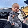 baby, infant, onesie, stars, grey_clothing, hand, truck, vehicle, outdoor, sky, cloudy, house, yellow_house, curious_expression, person, portrait, face, child, holding, casual