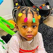 Anaelle a rejoint le concours — aidez-le/la à gagner de superbes lots ! toddler, child, braided_hair, colorful_hair_ties, indoor, rug, toy, stroller, curious, portrait, clothing, person, floor, expression, young_child, cute, sitting, face, eyes, playful