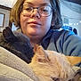 Stormy is registered to the contest to win money with this photo: person, glasses, cat, blanket, indoor, cozy, feline, face, portrait, short_hair, animal, snuggling, warm_lighting, home, furniture, comfort, resting, cute, pet, relaxation