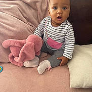 Manolie a rejoint le concours — aidez-le/la à gagner de superbes lots ! baby, child, infant, pink_blanket, stuffed_animal, soft_toy, striped_shirt, gray_pants, white_socks, couch, cushion, indoor, seated, expression, curious, wide_eyes, plush, home, cozy, portrait