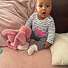 baby, child, infant, pink_blanket, stuffed_animal, soft_toy, striped_shirt, gray_pants, white_socks, couch, cushion, indoor, seated, expression, curious, wide_eyes, plush, home, cozy, portrait