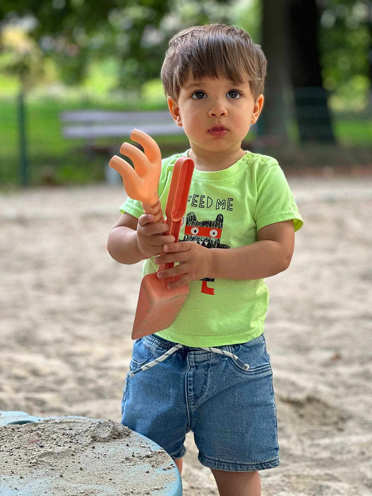 Liam participe au concours pour gagner de l'argent avec cette photo : child, toddler, sand, playground, toy, plastic_toy, green_tshirt, denim_shorts, outdoor, summer, cute, expression, person, holding, standing, nature, bench, sand_table, daylight, casual_clothing
