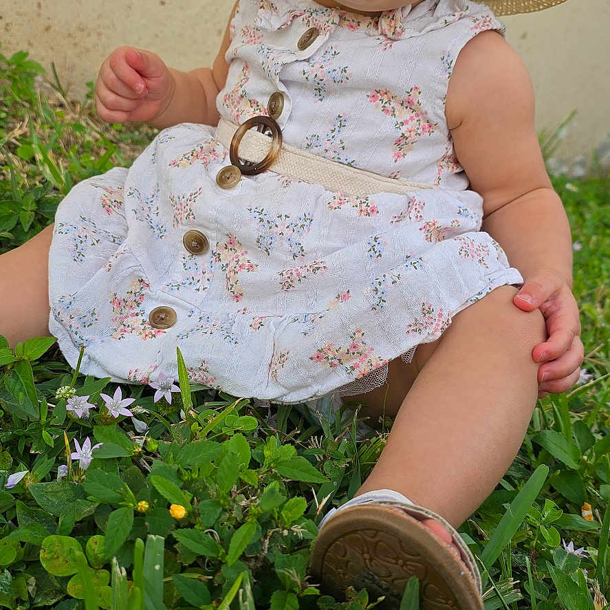 Amora is registered to the contest to win money with this photo: baby, clothing, dress, face, flower, footwear, grass, hat, head, herbal, herbs, lawn, outdoors, person, photography, plant, portrait, shoe, sitting, sunhat