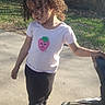 child, curly_hair, tshirt, strawberry_graphic, pants, sneakers, outdoor, sunny, sidewalk, stroller, smiling, playful, young, person, daylight, casual_clothing, nature, greenery, shadow, happy