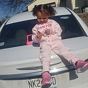 Brooklyn is registered to the contest to win money with this photo: toddler, child, car, white_car, pink_tracksuit, shoes, outdoor, daylight, smiling, playing, vehicle, license_plate, suburban, tree, house, sunny, shadow, person, happy, casual