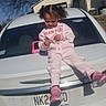 toddler, child, car, white_car, pink_tracksuit, shoes, outdoor, daylight, smiling, playing, vehicle, license_plate, suburban, tree, house, sunny, shadow, person, happy, casual