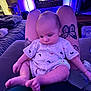 Grayson is registered to the contest to win money with this photo: baby, tattoo, legs, sitting, indoor, television, purple_light, onesie, foot, hand, person, furniture, couch, relaxation, home, nighttime, soft_lighting, casual, cute, infant