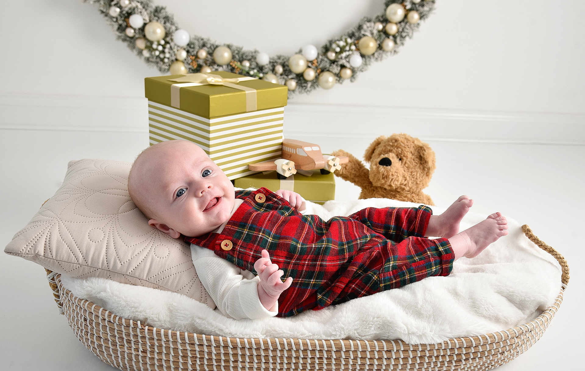 Leighton joined the competition — help win amazing prizes! baby, infant, plaid_overalls, smiling, basket, pillow, blanket, teddy_bear, toy_airplane, gift_box, holiday_wreath, decorations, indoor, cute, child, portrait, cozy, white_background, festive, happy