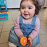 barefoot, child, curly_hair, cute, denim_dress, floor_tiles, foam_floor_tiles, hair_tie, happy, indoor, orange_toy, person, pink_shirt, plastic_chair, playful, sitting, smiling, toddler, toy, young_child