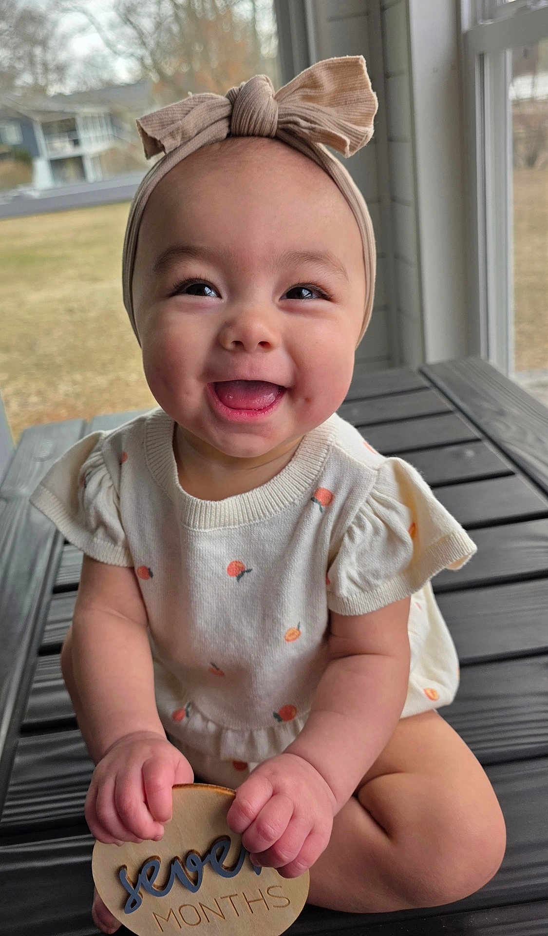 Rossi is registered to the contest to win money with this photo: baby, infant, smile, headband, onesie, milestone_disc, seven_months, hands, tongue, portrait, close_up, natural_light, porch, window, outdoor_background, cute, chubby_cheeks, sitting, wooden_toy, happy