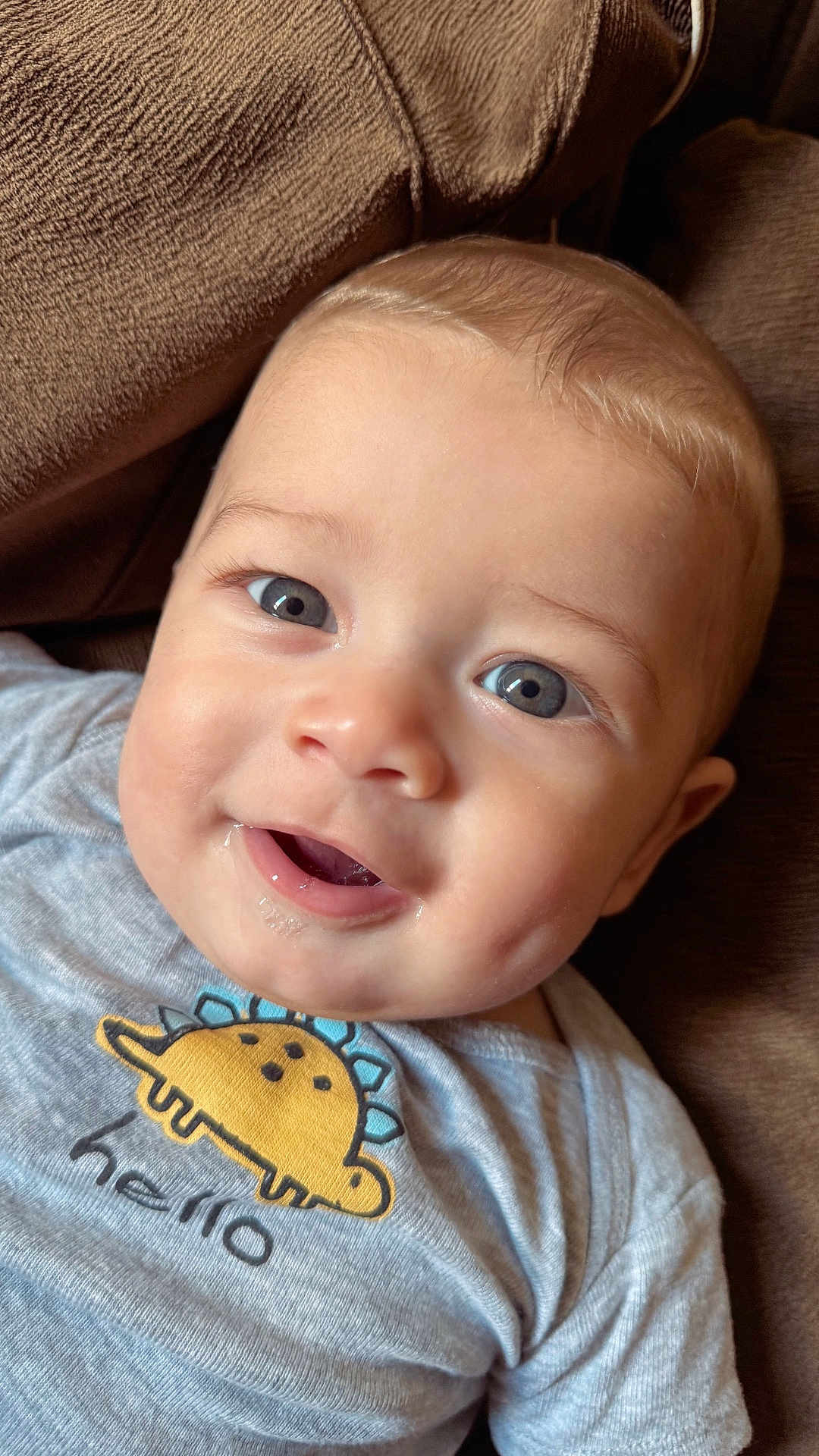 Leo is registered to the contest to win money with this photo: baby, child, smiling, face, blue_eyes, drool, dinosaur, shirt, hello, clothing, cute, close_up, infant, person, couch, brown, happy, indoors, portrait, young