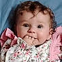 baby, child, curly_hair, face, fingers, floral_clothing, pink, portrait, cute, infant, lying_down, soft_texture, adorable, young_child, smiling_eyes, close_up, indoors, person, happy, toddler