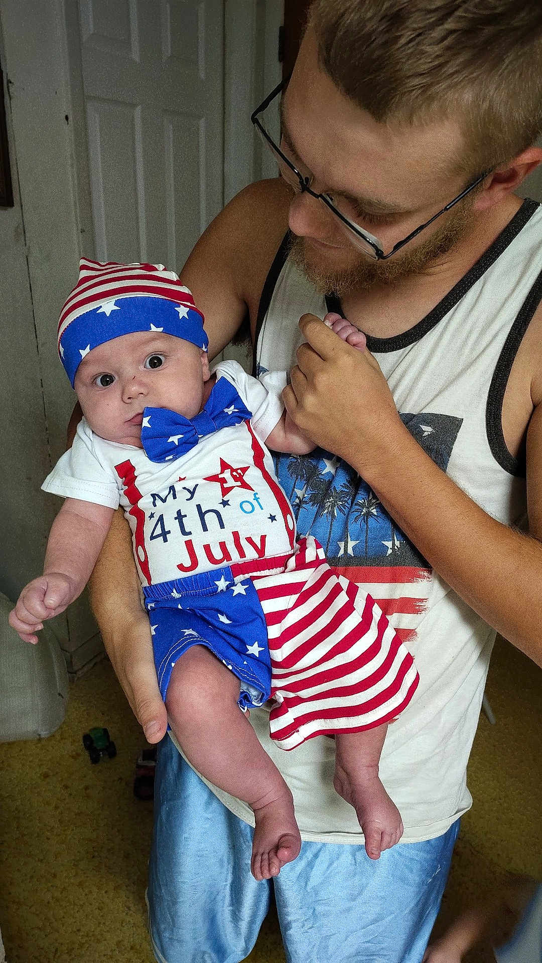 Apollo joined the competition — help win amazing prizes! baby, adult, holding, patriotic_clothing, 4th_of_july, stars_and_stripes, blue, red, white, hat, bowtie, glasses, facial_hair, tank_top, shorts, indoor, carpet, door, expression, person