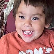 Romeo is registered to the contest to win money with this photo: child, toddler, smiling, happy, portrait, indoors, seated, orange_shirt, short_hair, brown_hair, teeth, open_mouth, face, head, eyes, chair, cushion, home, close_up, photograph