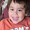 child, toddler, smiling, happy, portrait, indoors, seated, orange_shirt, short_hair, brown_hair, teeth, open_mouth, face, head, eyes, chair, cushion, home, close_up, photograph