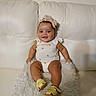 Aleyah joined the competition — help win amazing prizes! baby, infant, smiling, headband, yellow_sandals, white_onesie, couch, fluffy_cushion, happy, cute, indoors, child, seated, portrait, skin, person, furniture, legs, feet, comfort