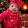 baby, infant, red_clothing, headwear, bow, christmas_tree, christmas_ornaments, gifts, wrapped_presents, fireplace, holiday, festive, indoors, smile, cozy, warm_lighting, face, celebration, cute, child