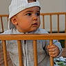 baby, backwards_cap, cap, cheeks, closeup, crib, cute, eyes, face, grip, hand, indoor, infant, neutral_expression, portrait, sitting, soft_lighting, sweater, toddler, wooden_rail