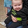 baby, child, toddler, infant, smiling, tongue_out, black_clothing, blue_shirt, adult_hand, sitting, indoor, floor, red_chair, jeans, sneakers, person, portrait, cute, happy, family