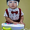 baby, child, helmet, bow_tie, red_suspender, toy, piano, sitting, curious, indoor, portrait, clothing, footwear, cute, young, infant, hands, face, expression, studio